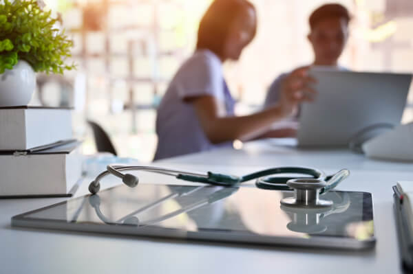 Zwei Pfleger schauen im Hintergrund zusammen in einen Bildschirm. Im Vordergrund liegt ein Stetoskop auf einem Tablet.