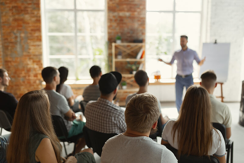 Das Bild zeigt einen Redner, der einen Vortrag vor einer Gruppe sitzender Menschen hält.