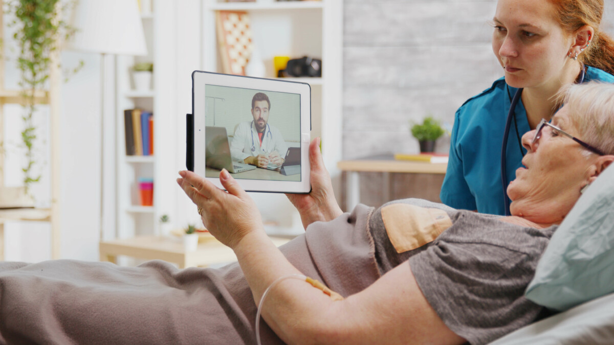 Das Bild zeigt eine Wohnsituation mit einer älteren pflegebedürftigen Dame, die im Bett liegt und ein Tablet hält, über welches sie sich in einer Videokonferenz mit einem Arzt unterhält. Eine jüngere Frau sitzt neben dem Bett und schaut ebenfalls in das Tablet.