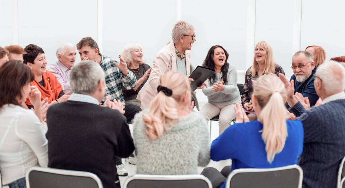 Das Bild zeigt eine Gruppe zumeist älterer Menschen, die im Kreis zusammensitzen und sich fröhlich miteinander austauschen.