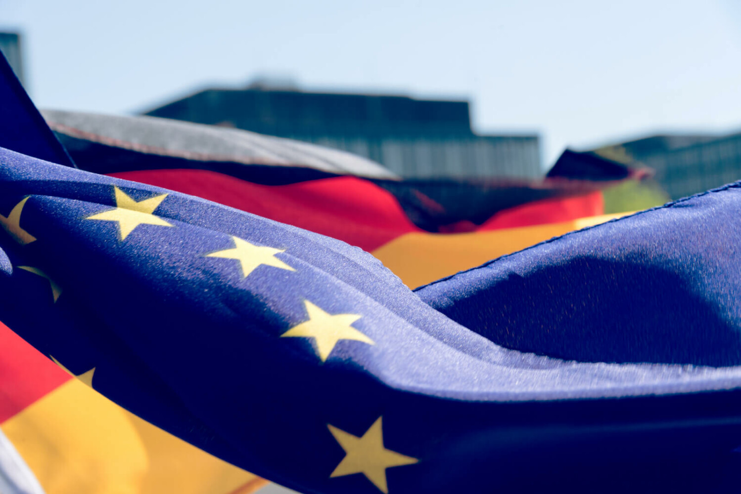 Europäische Flagge im Vordergrund dahinter die deutsche Flagge und verschwommen weiter weg der Reichstag in Berlin