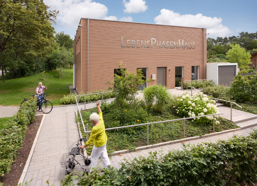 Eine ältere Frau mit Rollator grüßt einen älteren Mann mit Fahrrad. Beide stehen vor einem Haus mit der Aufschrift Lebensphasenhaus.