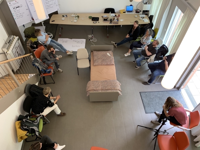 Das Bild ist im LebensPhasenHaus aufgenommen. Es zeigt eine Gruppe von Menschen, die sich während eines Workshops sitzend um ein Pflegebett gruppiert haben und sich über technische Neuerungen in der Pflege austauschen.